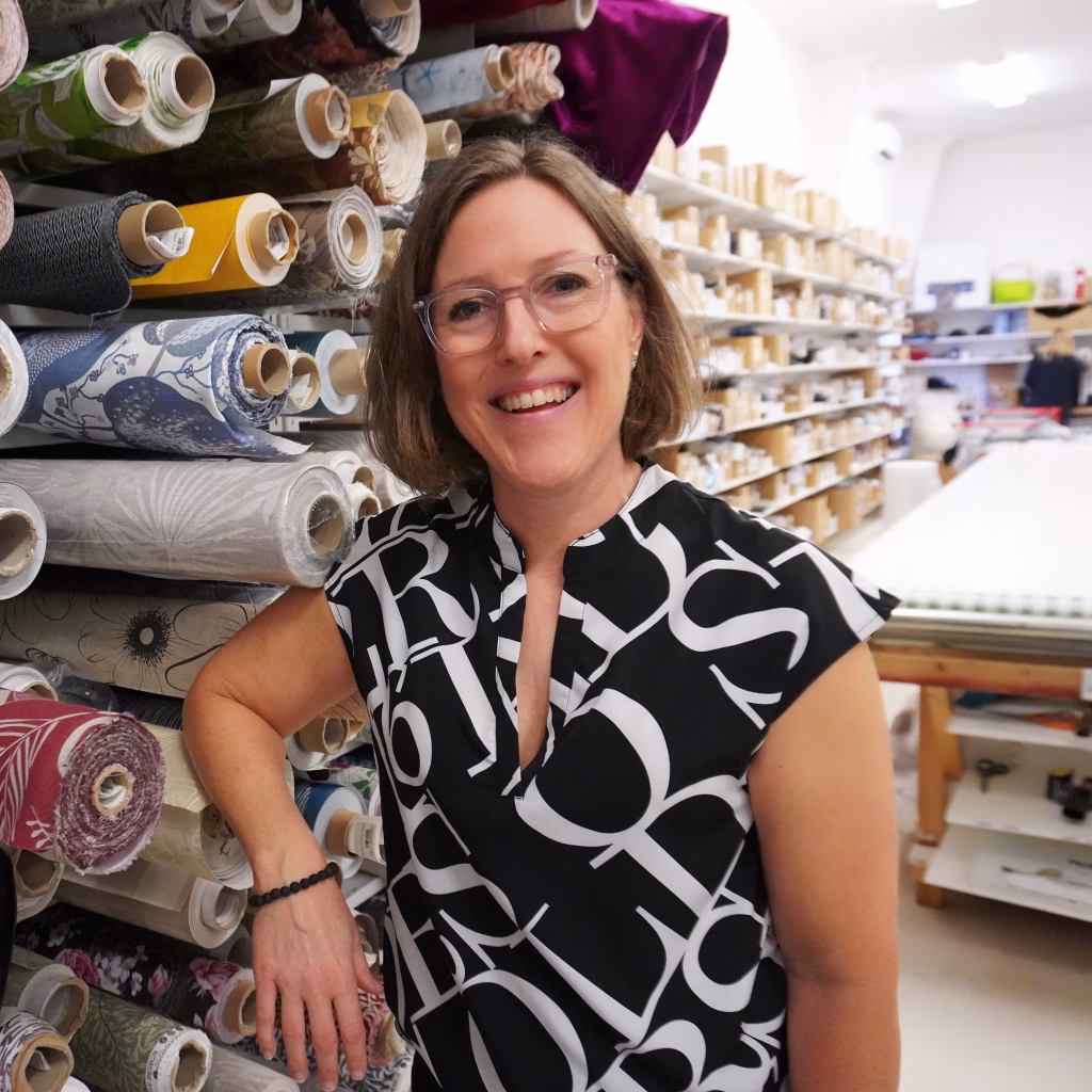 En leende kvinna står intill en vägg fylld med hyllplan med rullar av tyger på.