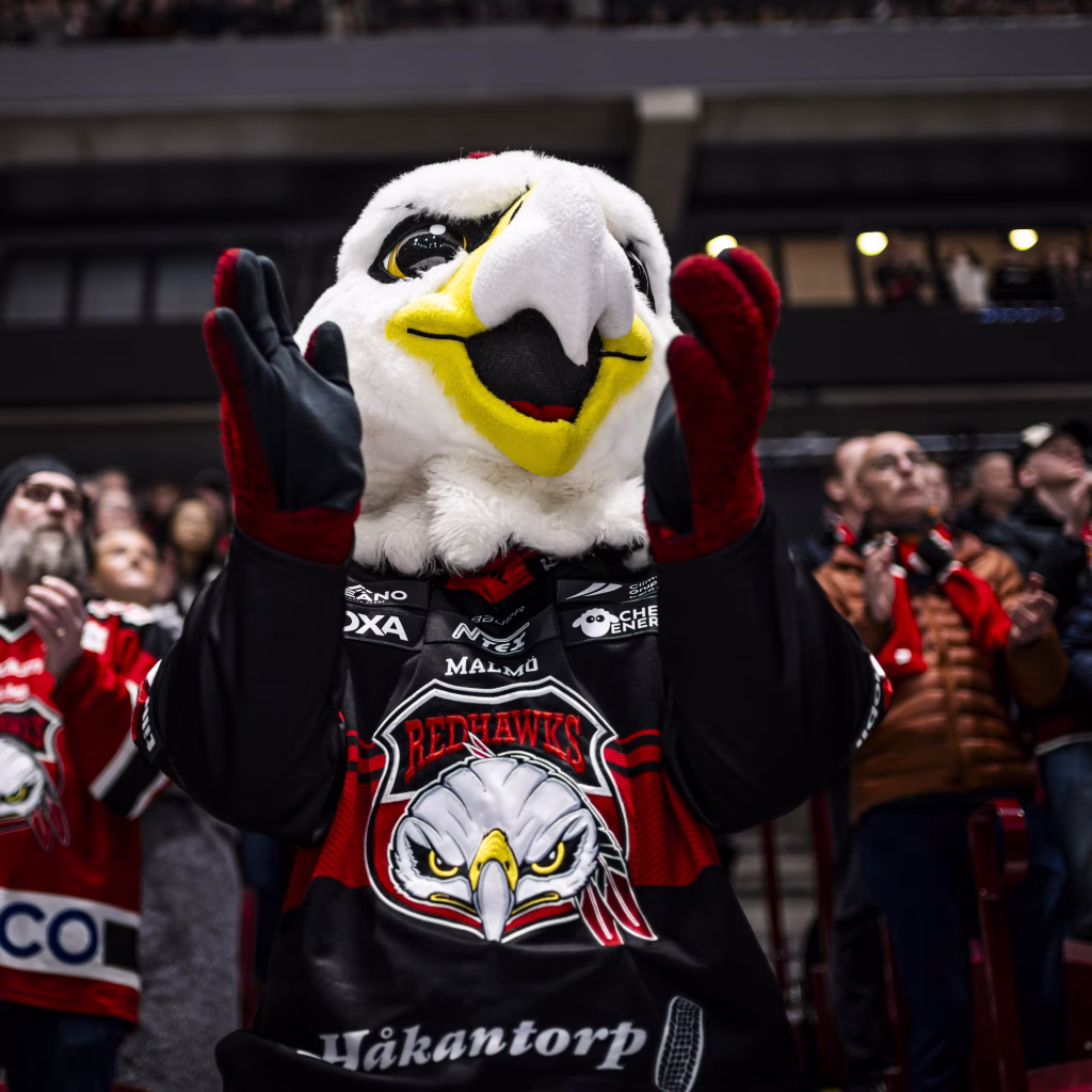 Malmö Redhawks maskot "Hawkan" står på läktaren och klappar. I bakgrunden är Malmö Redhawks supportrar som också står på läktaren och klappar.