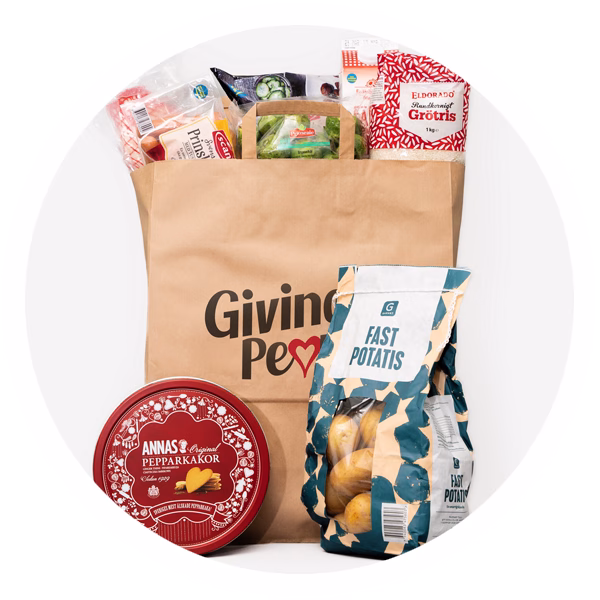 En papperskasse fylld med matvaror. Papperskassen har Giving People logotypen på. Ur kassen sticker paket med grötris, brysselkål, köttbullar och prinskorv halvt ut. Utanför påsen står en förpackning av Annas Pepparkakor och en två kilo förpackning av Garants fast potatis.